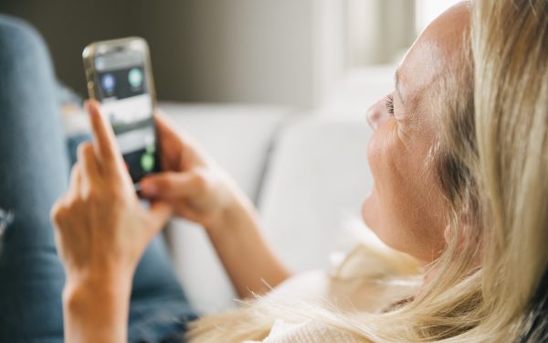 En dame med blondt hår som ligger på sofaen og smiler og holder en mobiltelen med en app-løsning fra porttelefon-leverandøren Urmet